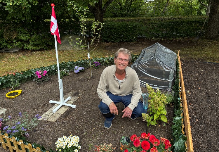 Max på hug i sit havelod i Gyvelparken Ry. Kigger op og smiler til kameraet omringet af et flag, blomster og drivhus. Foto: Skanderborg Kommune